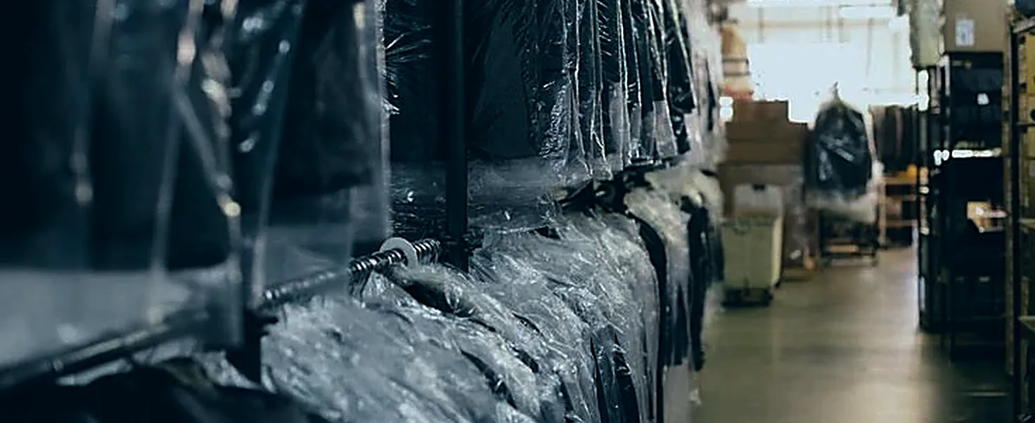 Finished garments hanging in protective covers along a dry cleaning rack, an aisle receding towards daylight
